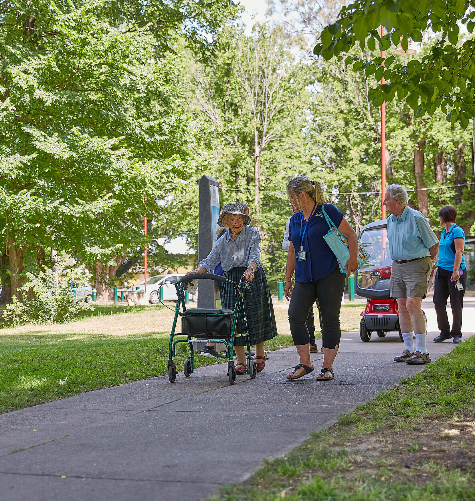 A woman in a long green skirt and blue shirt walking down a garden path using a walker. There are bright green trees in the background and another Woman in a Valmar uniform is walking beside her.
