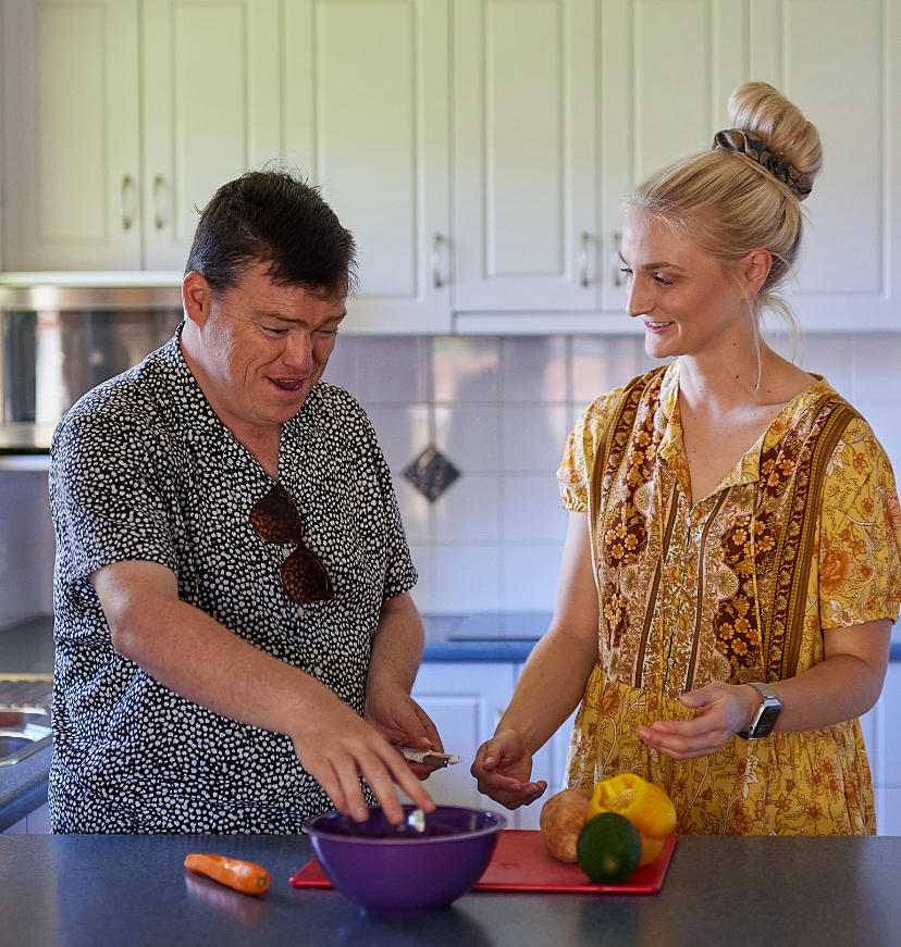 A young woman and man at a kitchen bench, he is a paisley shirt and she is wearing a yellow dress. They are cutting vegetables on a red chopping board and placing them in a purple bowl.