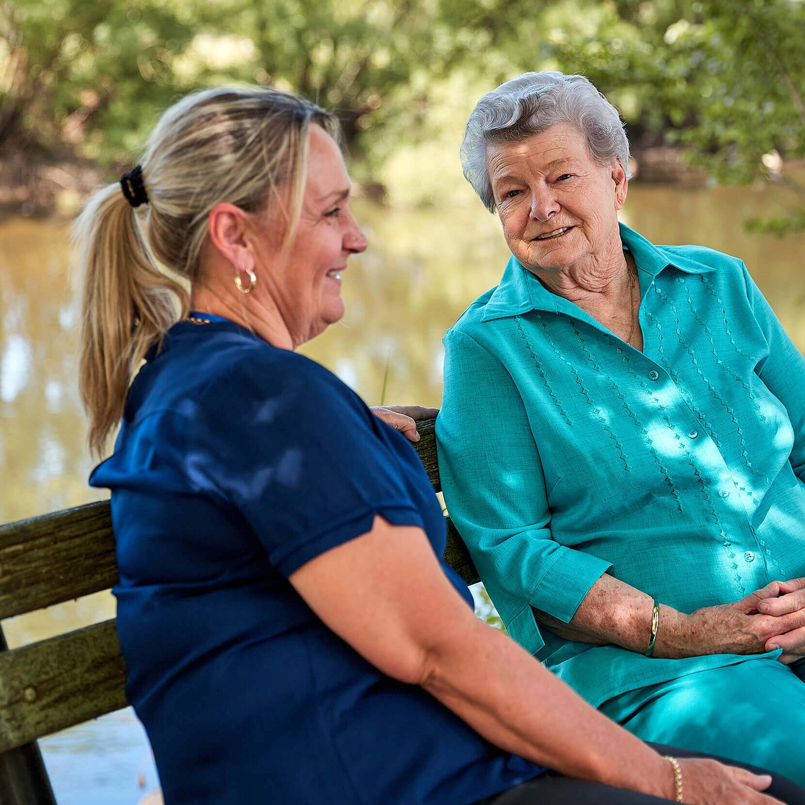 A blonde woman sitting on a park bench smiling in a blue Valmar uniform. An elderly woman in a green pant suit is sitting next to her with her hands crossed in her lap looking happy.