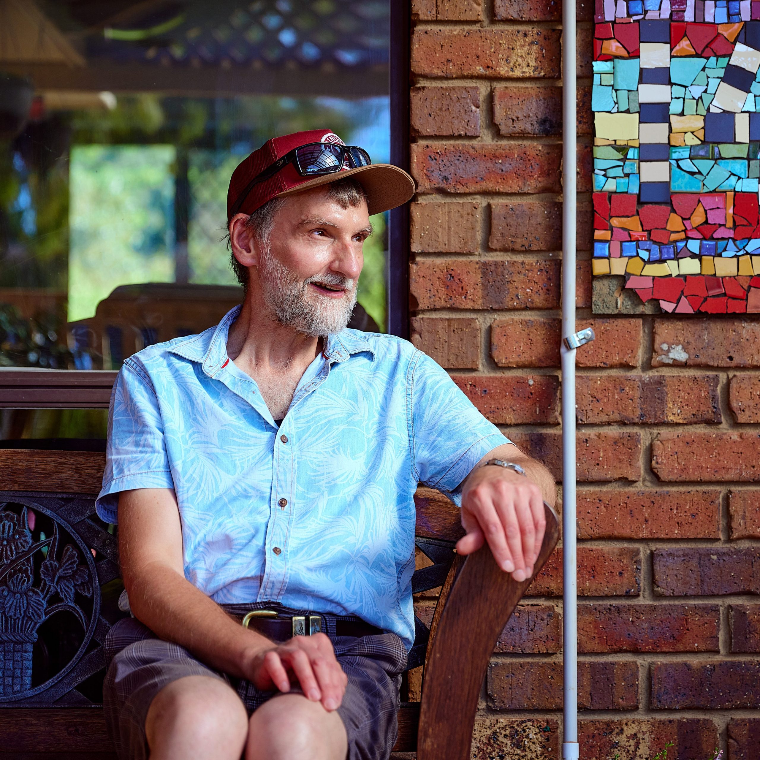A man smiling in a light blue shirt and dark grey shorts sitting on a bench on the porch of his home
